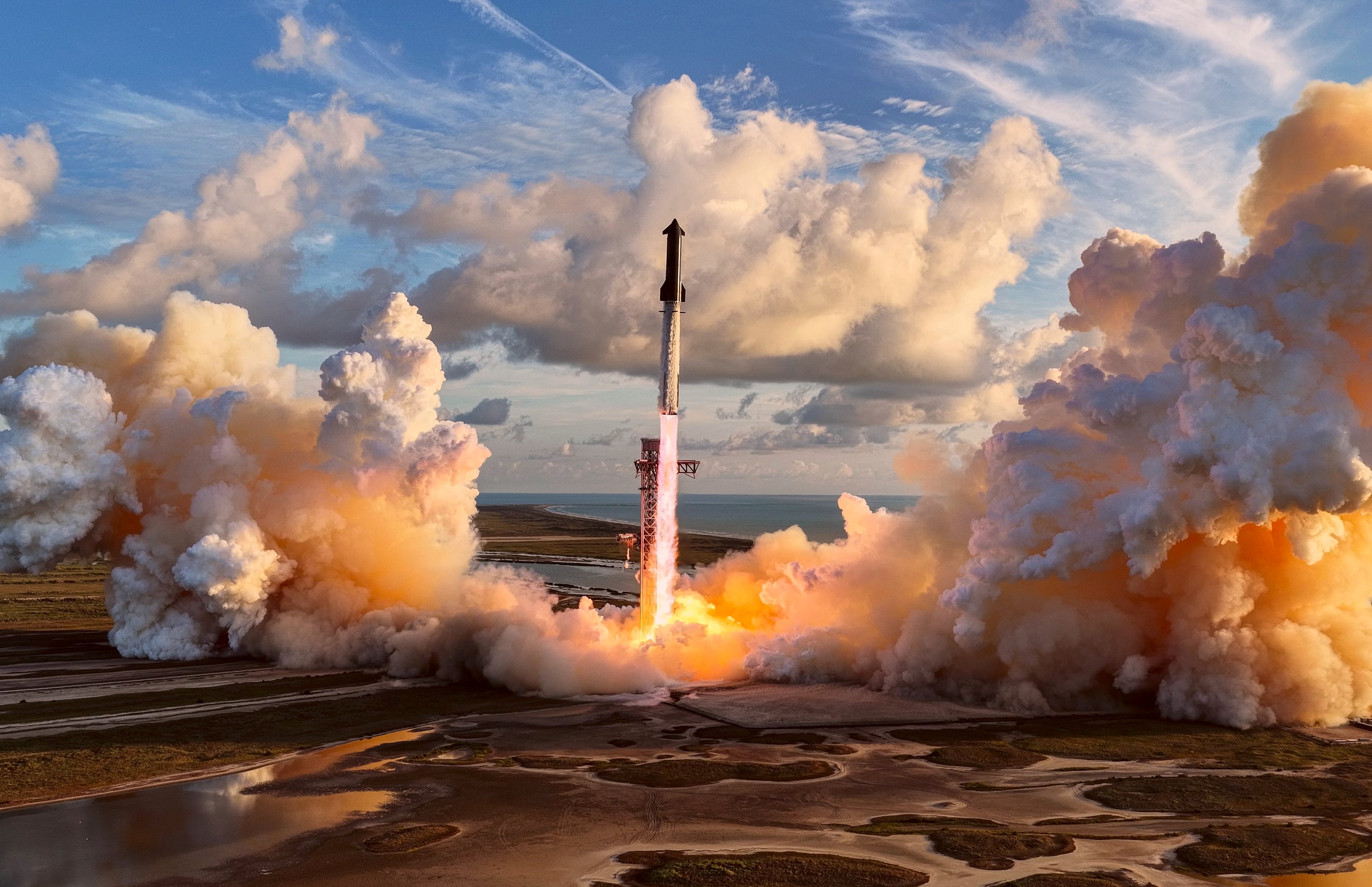 SpaceX Starship lifting off through orange-lit smoke beneath a blue sky